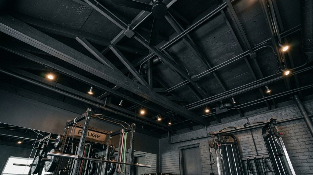 Modern industrial gym interior featuring a black painted ceiling with exposed wooden beams, conduits, and warm spotlights.