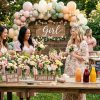 Three women arrange pastel flowers at a garden baby shower 'Flower Bar' featuring a balloon arch and mocktail station.