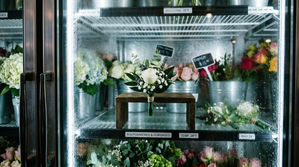 A delicate white rose boutonniere sits on a wooden stand inside a floral refrigerator filled with colorful flowers.